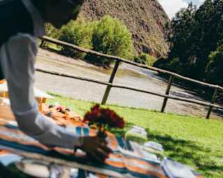 In soft-focus foreground, a waitress places a budvase on an dining table on the banks of the Urubamba, shot at an angle.
