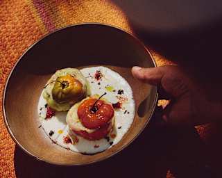 A hand holds a brown bowl with two stuffed vegetables, one red and one green, on a creamy white sauce, garnished with herbs and spices, placed on a textured orange surface.