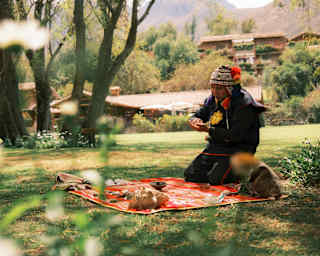 A shaman in a traditional chullo hat kneels on a red-patterned blanket laid on the lawn and covered with shells and stones.