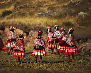 Women gather on a grassed area, dressed in vibrant traditional skirts, Manta shawls and Montera hats with sanq'apa straps.