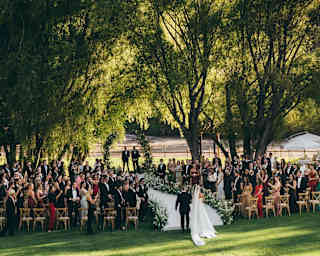 Guests cheer as a bride and suited man arrive at an aisle, set beneath shady garden trees, with a dramatic mountain backdrop.