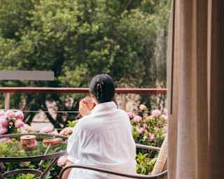 A woman with dark hair, wearing a white robe, sips from a cup as she sits on a deck gazing at the scenery, seen from behind.