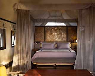 View over a table to a double bed against a partition wall, with side tables and lamps, and a canopy with tied-back drapes.