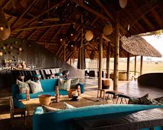 Turquoise sofas rest beneath the thatched roof of a deck with open sides to views of grasslands, with an elephant in sight.