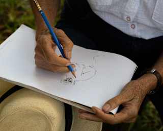 Close-up of Vicki Thomas's hands as she sketches a monkey with a blue pencil, with her straw hat hanging on one knee.
