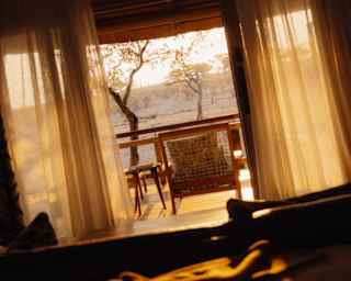 Angled image of a deck overlooking the plains, bathed in golden sun, seen through open drapes from a lodge interior.