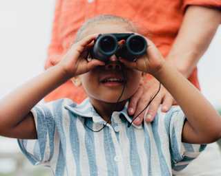 A man in an orange shirt rests his hand on the shoulder of a young boy in a blue striped top, who looks through binoculars.