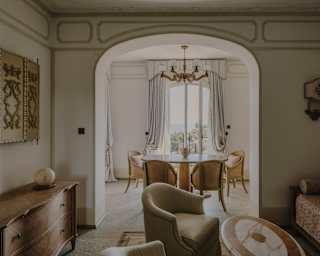 View through the Riviera Suite living area and wall arch to a dining table with cane chairs by a tall window with white drapes.