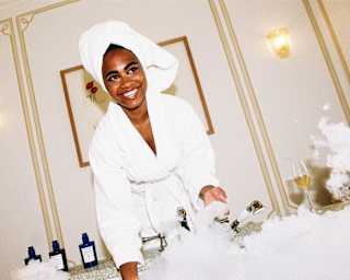 Angled image of a woman in a white robe, hair wrapped in a towel and smiling as she puts a hand in a luxurious bubble bath.