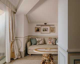 A beige sofa with floral cushions is tucked beneath a gabled ceiling in the Junior Suite, seen over an ornate pedestal table.