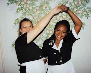 Two female staff members in black and white uniforms smile as as they twirl each other in a dance in front of a floral mural.