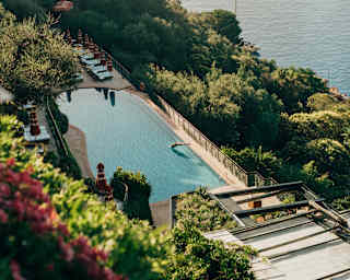 Action shot as a guest is captured mid-air diving into the glass-like pool, in a view from above over gardens and sea.