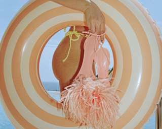 A woman in a bikini holds an blow-up ring under an arm, and pink, Bottega Veneta-like feather sandals in her hand, close-up.