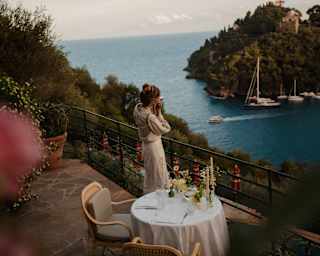 Seen from above, a woman in white takes photographs of Baia Cannone from the terrace where a dining table is laid for two.