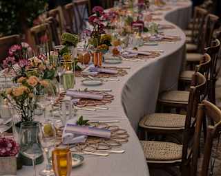 Close-up of a wavy banquet table adorned with pretty flower vases and place settings with wicker mats, napkins and scrolls.