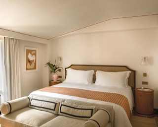 Bright hotel room with a large bed, white linen, tan and orange accents, a small sofa at the foot of the bed, artwork on the wall, a window with open curtains, and bedside tables.