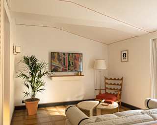 A cosy living room with a potted plant, wooden chair with red cushion, round table, sofa, wall art, and warm lighting. A large window lets in natural light.