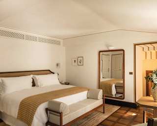 A cosy hotel room with a large bed, beige bedding, a bench at the foot, a big mirror reflecting the space, a table with a floral vase, and soft natural lighting. The decor is modern and minimalist.