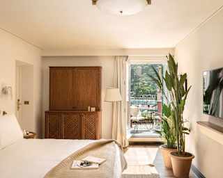 Sunbathed bedroom with a bed, armoire, live plants and an open door leading to the balcony