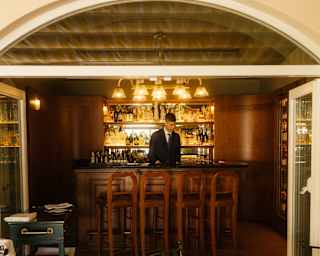 A barman stands behind a wood-panelled bar below a glowing chandelier, in front of backlit bottle shelves, seen from outside.
