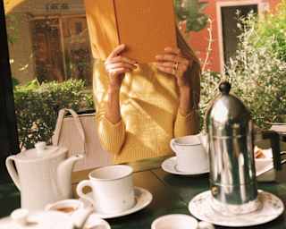 A woman in a yellow top holds an orange menu book in front of her face, seen over a table covered with coffee and teacups.