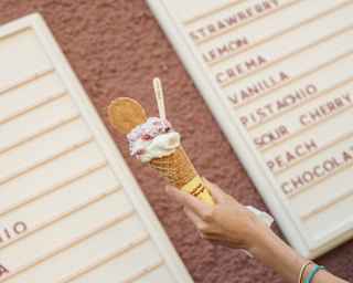 Angled image of woman's hand as she holds up an ice-cream cone with a wafer against a backdrop of two gelateria menu boards.