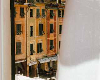 View from a high part-draped window down on to La Piazzetta's peach pastel buildings and an eatery's outdoor seating.