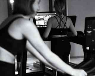 Monochrome image of one woman side-on in soft-focus foreground and another from behind, as they exercise on gym machines.