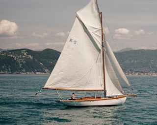 A vintage K-2 sailing yacht, with white hull and sails, cruises across the azure Ligurian waters, skippered by a single man.