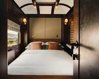 View into a double cabin with an immaculate white bed topped with russet cushions, flanked by lamps, seen through a doorway.