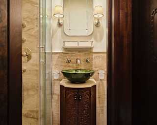 A green basin rests on a carved wooden vanity with a wall mirror, flanked by vintage lamps, in a marble-tiled bathroom.