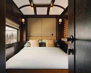 A double bed fills the polished wood cabin, with white linen, cream cushions and glowing wall lamps, seen through an open door.