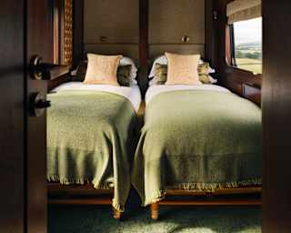 Two fold-down beds, made up with white linen and olive-green blankets, seen in a wood-panelled twin cabin from an open door.