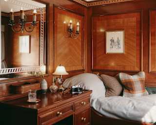 View of a wooden desk perpendicular to a bed with white bedding and cushions, in a panelled twin cabin with wall lamps.