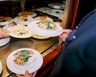 A waiter lifts two plates from the pass, both containing two juicy pan-fried scallops with herb crust, served in the shell.