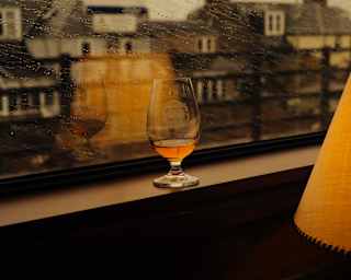A Glencairn tasting whisky glass containing an amber nip rests on the sill of a rain-spattered window next to a glowing lamp.