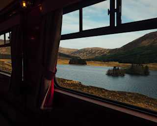 The train passes a loch with a forested island and mountainous backdrop in bronze and heather, seen through a window.