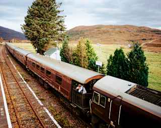 A man looks out from an observation deck as The Royal Scotsman waits at a rural station in the Highlands, seen from above.