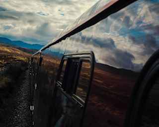 Close-up view down the exterior of the train, its carriages so glossy that they mirror the cloudy sky and the rugged highlands.