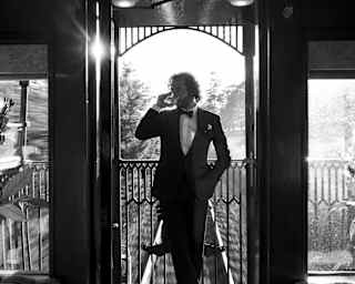 In black and white, a male guest in black-tie sips a drink with one hand in his pocket on the open-air observation deck.