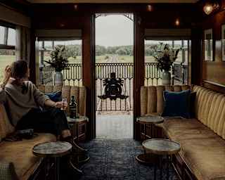 A woman relaxes with a drink on a banquette of warm gold upholstery as she gazes through the door of the observation deck.