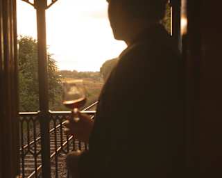 A man holding a drink, seen in half-silhouette, stands on the Observation Car deck, gazing back at the tracks at sundown.