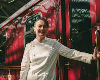 Chef Lorna McNee stands by the cherry exterior of The Royal Scotsman with one hand on a door handle, in a low-angle close-up.