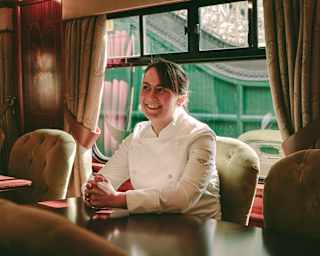 Chef Lorna McNee, wearing chef whites, sits behind a table inside a restaurant car with her hands clasped in front of her, smiling.