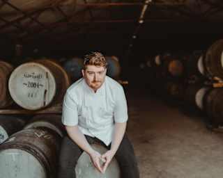 Chef Mark Donald, with red hair and beard, sits on a barrel in the Glenturret Distillery wearing chef whites and dark trousers.