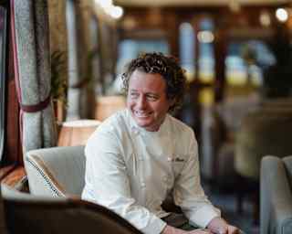 Portrait of Chef Tom Kitchin in his chef whites, as he gazes out of a window with a soft smile, seated in a blue armchair.