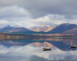 Two sailing boats sit on the glasslike waters of Loch Eil which mirror the surrounds of green fields and snow-capped mountains.