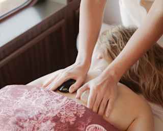 A spa therapist holds massage stones on the back of a woman covered by a burgundy Toile De Jouy sheet, at the Spa by Dior.