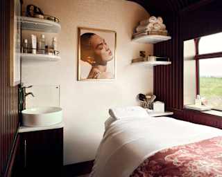 View into a spa room with corner shelves and a bed with white sheets and Dior's trademark Toile de Jouy throw in mauve.