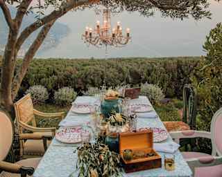 The garden table is set for a celebration beneath a chandelier suspended from an olive tree, with breathtaking coastal views.